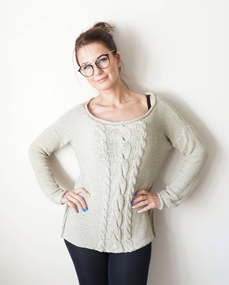 May include: A woman wearing a light beige cable knit sweater with long sleeves. She is wearing black pants and has her hair pulled back in a bun. She is wearing black framed glasses and has blue nail polish.