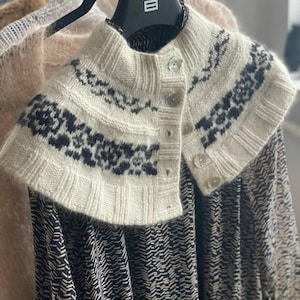Könnte beinhalten: Ein weißer Strickkragen mit einem schwarzen geometrischen Muster und drei weißen Knöpfen. Der Kragen wird über einem schwarz-weiß gemusterten Kleid getragen.
