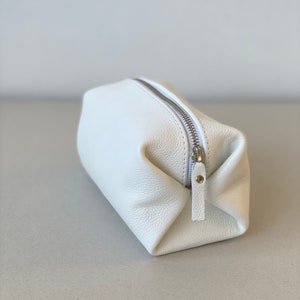 May include: A white leather zippered pouch or cosmetic bag. The bag is folded and has a silver zipper and pull.