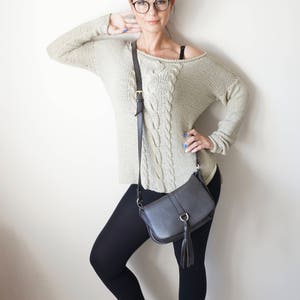 May include: A woman wearing a light beige cable knit sweater, black leggings, white sneakers, and a black crossbody bag with a tassel.