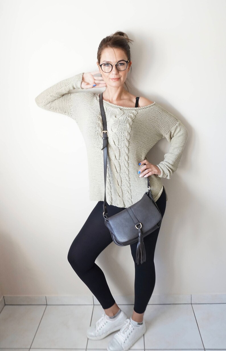 May include: A woman wearing a light beige cable knit sweater, black leggings, white sneakers, and a black crossbody bag with a tassel.
