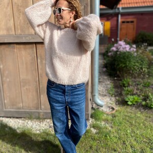 Puede incluir: Una mujer con un su&eacute;ter de color rosa claro y peludo de corte holgado y jeans azules. Est&aacute; de pie frente a una pared de madera y tiene el pelo recogido hacia atr&aacute;s.