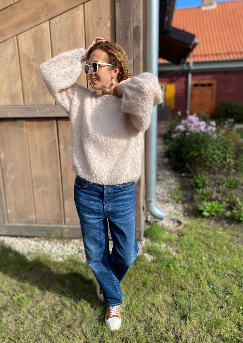Puede incluir: Una mujer con un su&eacute;ter de color rosa claro y peludo de corte holgado y jeans azules. Est&aacute; de pie frente a una pared de madera y tiene el pelo recogido hacia atr&aacute;s.