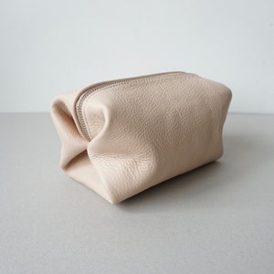 May include: A light beige leather zippered pouch or cosmetic bag. The bag is folded and sitting on a white surface.