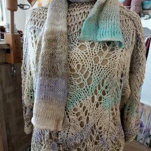 Peut inclure: Un ensemble tricoté à la main, composé d'un pull ajouré et d'une écharpe. Le pull est beige avec des touches turquoise et lavande. L'écharpe présente un dégradé de couleurs, dont le gris, le lavande et le turquoise. L'étiquette de l'écharpe indique "AQUAREVI".