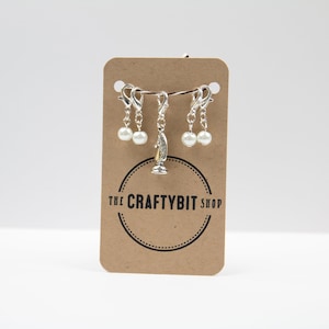 Op de afbeelding: Een set van vier zilveren steekmarkeerders met witte parel kralen en een zilveren schaakstuk bedel. De markeerders worden weergegeven op een bruine kaart met de tekst "The Craftybit Shop".