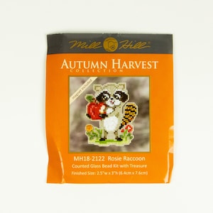 May include: A close-up of a Mill Hill Autumn Harvest Collection counted glass bead kit featuring a raccoon holding an apple. The kit is called "Rosie Raccoon" and the finished size is 2.5 inches wide by 3 inches high (6.4 cm x 7.6 cm).