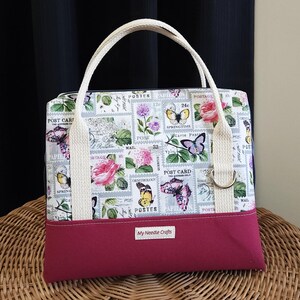 May include: A small, white tote bag with a red base. The bag is decorated with a vintage postage stamp pattern featuring butterflies and flowers. The text "My Needle Crafts" is printed on the bottom of the bag.