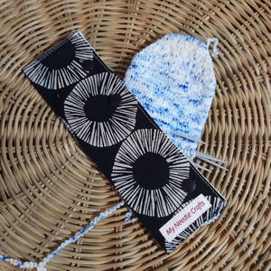 Peut inclure: Un étui à aiguilles noir et blanc avec un motif circulaire et le texte "My Needle Crafts" est affiché à côté d'un article tricoté bleu et blanc. Les articles sont sur une surface en osier tressé.