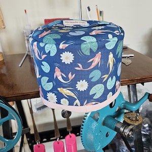 May include: A blue and white fabric yarn bowl with a pink trim and a koi fish and lily pad pattern. The bowl is on a yarn winder with a turquoise wheel and a wooden handle. The text "My Needle Crafts" is on the bowl.