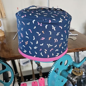 May include: A blue fabric cover with a pink trim and a white and orange confetti pattern. The cover is on a spinning wheel with a teal gear and a black handle.
