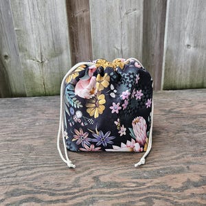 May include: A black drawstring bag with a colourful floral design, including pink, yellow, and purple flowers. The bag has a white drawstring closure and is made of fabric. The background is a wooden surface.