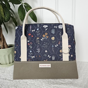 May include: A navy blue fabric bag featuring a white hexagonal pattern with various floral illustrations. The bag has cream-coloured canvas handles, a solid taupe base, and a small white label with the text My Needle Crafts.