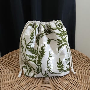 May include: A drawstring bag made of natural linen fabric with a green floral pattern. The bag features a white floral design with green leaves.