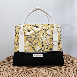 May include: A rectangular tote bag with a floral and bee pattern on a yellow background. The bag has a black base, cream-colored handles, and a metal ring detail. The text "My Needle Crafts" is visible on the front.