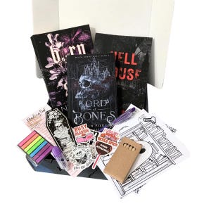 May include: A gift box filled with books, stickers, and stationery. The books have dark covers with gothic themes and titles like "Lord of Bones" and "Hell House." Other items include a pen, sticky notes, and a coloring page.