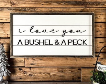 May include: A white wooden sign with black text that reads "i love you a bushel & a peck". The sign is hanging on a wood wall with a small white tree and a metal tiered stand with green plants and brown fruit on the bottom tier.