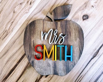 May include: Wooden apple-shaped sign with the text "Mrs. Smith" in rainbow-colored letters. The letters are raised and attached to the sign.