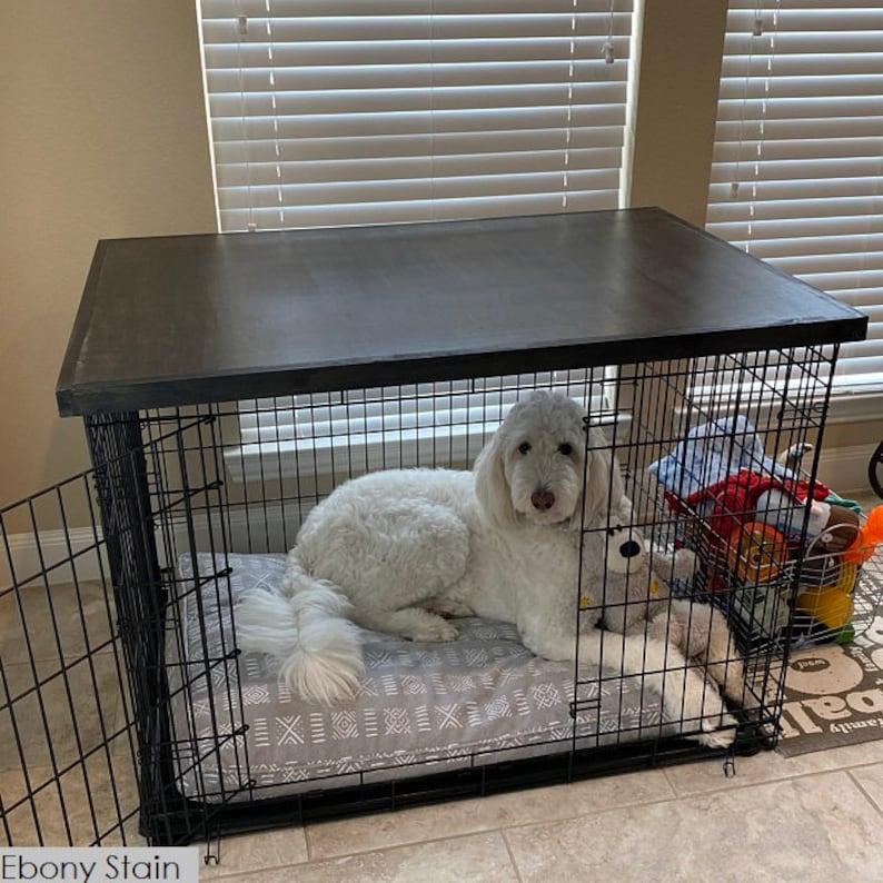 May include: A white dog is lying in a black metal dog crate with a dark gray wooden top. The crate has a gray and white patterned dog bed inside. The text "Ebony Stain" is visible at the bottom of the image.