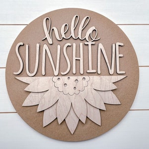 Pode incluir: Um sinal redondo de madeira com as palavras "hello sunshine" em uma fonte decorativa. O sinal tem um design de girassol em camadas no centro.