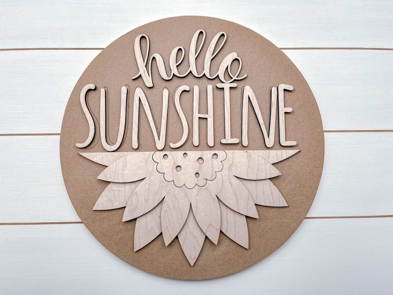 Pode incluir: Um sinal redondo de madeira com as palavras "hello sunshine" em uma fonte decorativa. O sinal tem um design de girassol em camadas no centro.