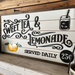 Op de afbeelding: Een wit houten bord met zwarte letters waarop staat "Sweet Tea & Lemonade Served Daily 25&cent;". Het bord heeft een zwart silhouet van een glazen pot met een rietje en ijsblokjes. Er zijn twee gele citroenen met groene bladeren op het bord.