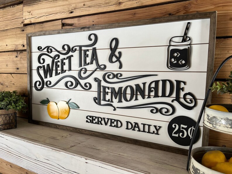Op de afbeelding: Een wit houten bord met zwarte letters waarop staat "Sweet Tea & Lemonade Served Daily 25&cent;". Het bord heeft een zwart silhouet van een glazen pot met een rietje en ijsblokjes. Er zijn twee gele citroenen met groene bladeren op het bord.