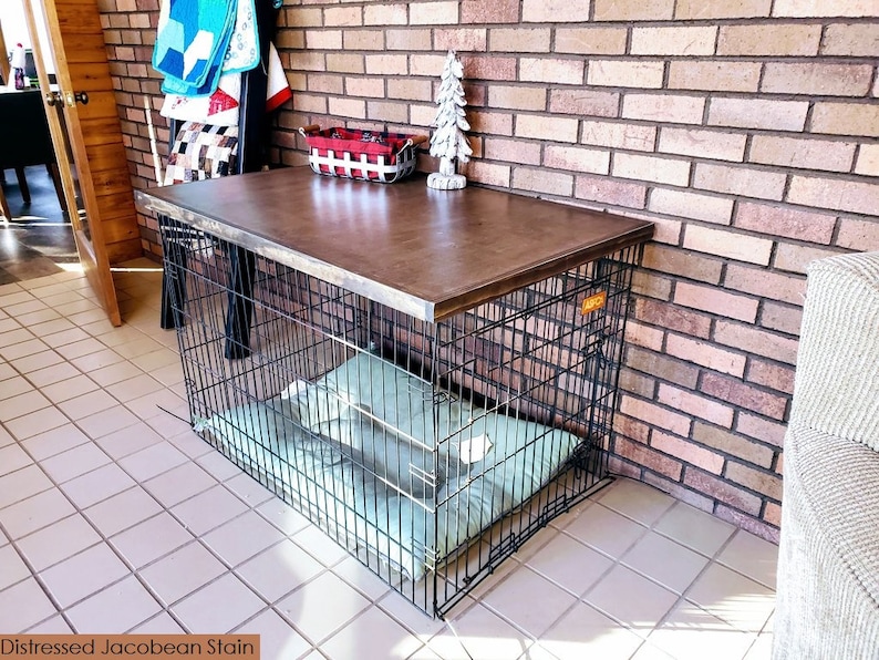 May include: A black metal dog crate with a distressed Jacobean stained wood top. The crate is sitting on a tiled floor in front of a brick wall. A green dog bed is inside the crate.