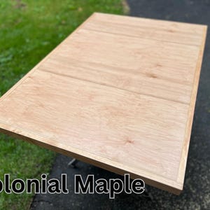 May include: A close-up of a light wood table top with a natural wood grain pattern. The table top has a slightly raised edge around the perimeter. The text "Colonial Maple" is printed below the table top.