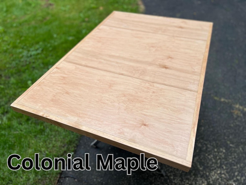 May include: A close-up of a light wood table top with a natural wood grain pattern. The table top has a slightly raised edge around the perimeter. The text "Colonial Maple" is printed below the table top.