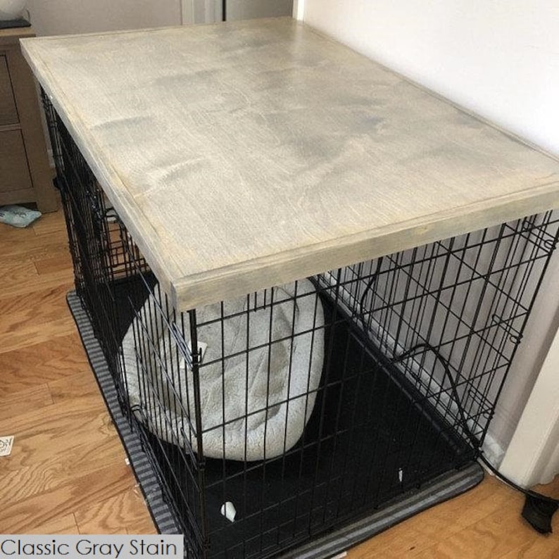 May include: A black metal dog crate with a gray wooden top. The crate has a black and white striped mat inside and a gray dog bed.