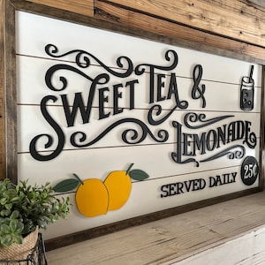 Op de afbeelding: Een houten bord met de tekst "Sweet Tea & Lemonade Served Daily 25&cent;" in zwarte letters. Het bord is versierd met citroenen en een kan limonade.