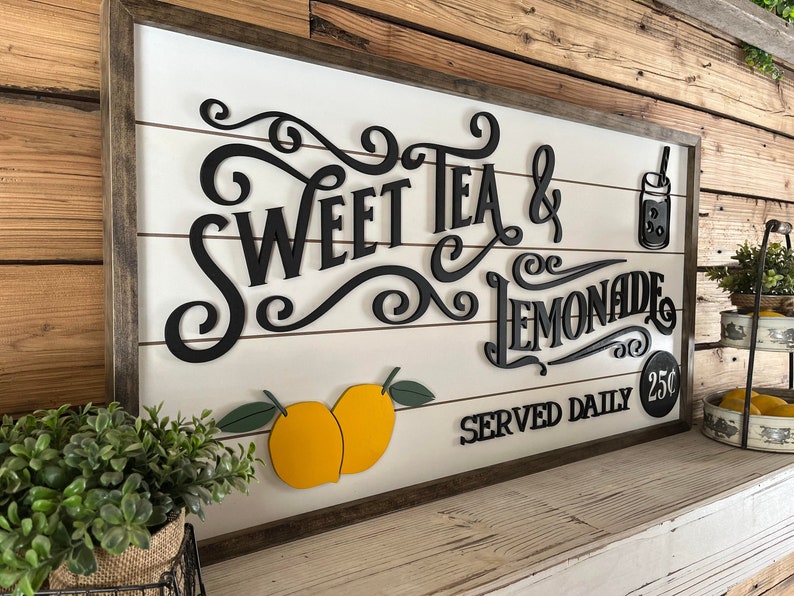 Op de afbeelding: Een houten bord met de tekst "Sweet Tea & Lemonade Served Daily 25&cent;" in zwarte letters. Het bord is versierd met citroenen en een kan limonade.