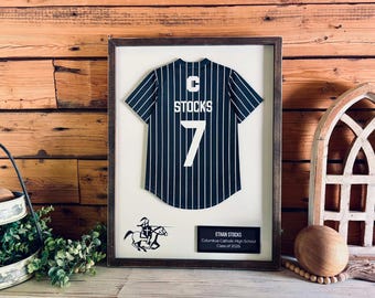 Pode incluir: Uma moldura exibindo uma camisa de beisebol azul-marinho com riscas, com a letra C, o nome STOCKS e o número 7 em branco. Abaixo da camisa, há um gráfico de um cavaleiro em um cavalo e uma placa preta com os dizeres ETHAN STOCKS, Columbus Catholic High School, Class of 2026.