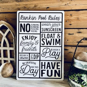 May include: White metal sign with black text that reads "Rankin Pool Rules". The sign has a grid of boxes with different rules for pool behavior, including "No Dunking, Running, Pushing, Peeing", "Wear Your Sunscreen", "Enjoy Family & Friends", "Float & Swim", "Relax & Unwind", "Sip & Dip", "Laugh & Play", "Be Grateful For This Day", and "Have Fun".