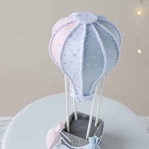 May include: A blue and pink hot air balloon cake topper with white stars and a rope design. The balloon is attached to a gray basket with two small bags hanging from it.