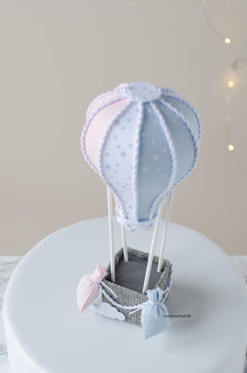 May include: A blue and pink hot air balloon cake topper with white stars and a rope design. The balloon is attached to a gray basket with two small bags hanging from it.