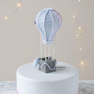 May include: A white cake with a blue and pink ribbon around the bottom. A blue and white hot air balloon with a gray basket sits on top of the cake.