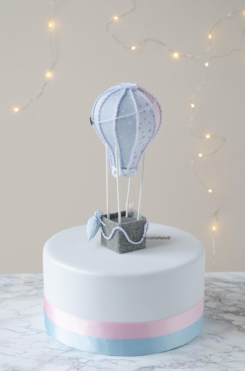 May include: A white cake with a blue and pink ribbon around the bottom. A blue and white hot air balloon with a gray basket sits on top of the cake.