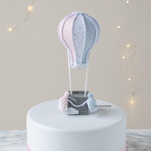 May include: A white cake with a blue and pink ribbon around the bottom. On top of the cake is a blue and pink hot air balloon with a gray basket and a white cloud.
