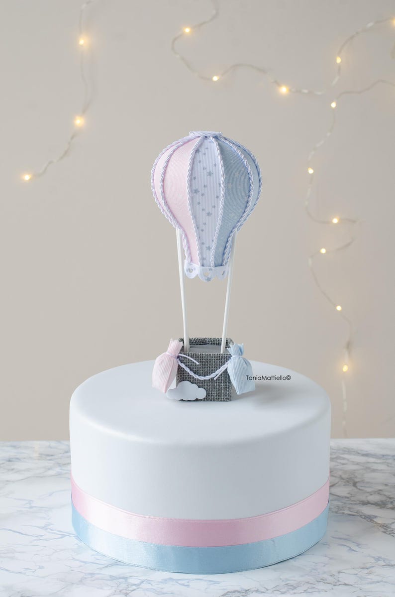May include: A white cake with a blue and pink ribbon around the bottom. On top of the cake is a blue and pink hot air balloon with a gray basket and a white cloud.