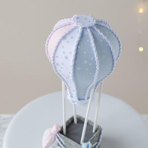 May include: A blue and pink hot air balloon with white stars and a gray basket. The balloon is decorated with white rope and has two small bags hanging from the basket. The text "TaniaMattiello&reg;" is visible at the bottom of the image.