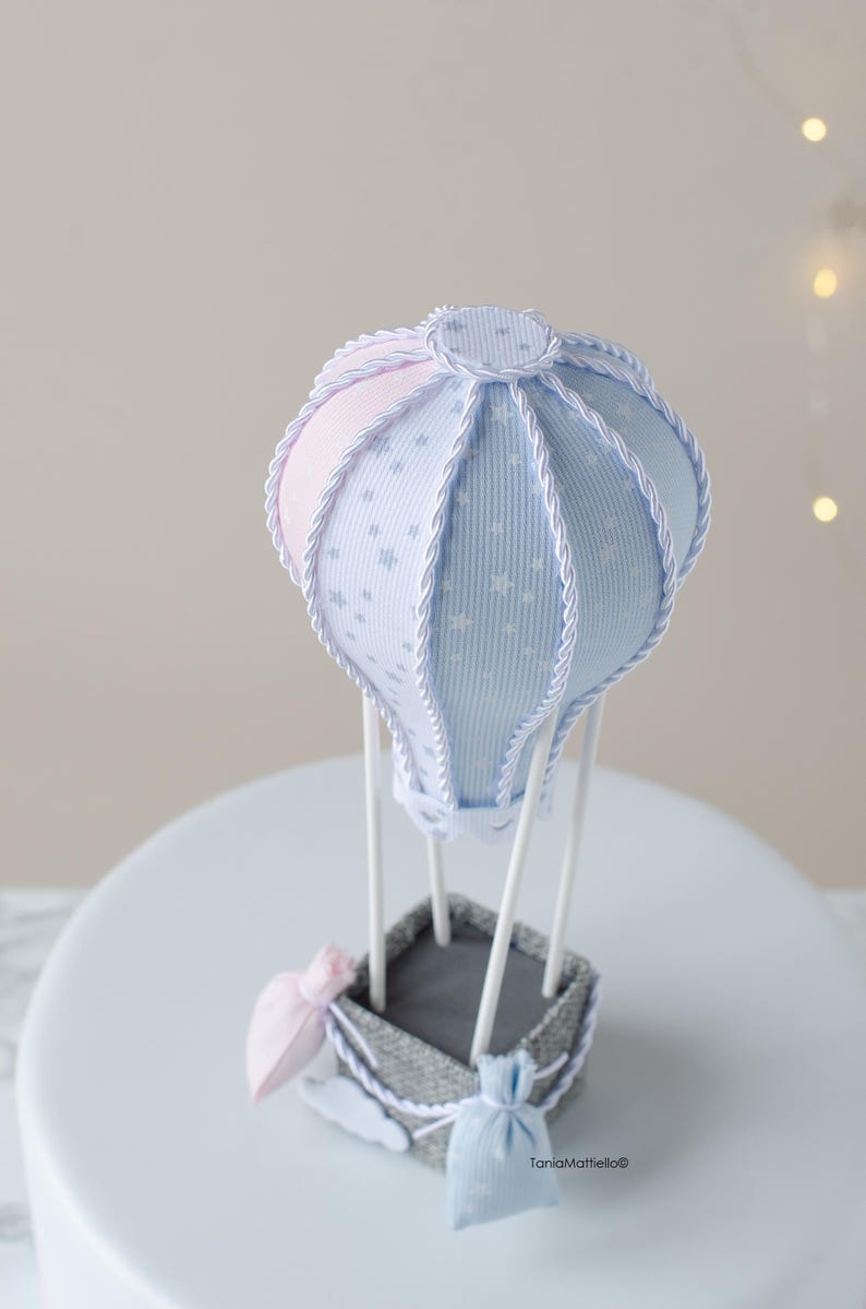 May include: A blue and pink hot air balloon with white stars and a gray basket. The balloon is decorated with white rope and has two small bags hanging from the basket. The text "TaniaMattiello&reg;" is visible at the bottom of the image.