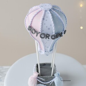 May include: A gender reveal hot air balloon cake topper with a pink and blue striped balloon, white stars, and the text "Boy or Girl!" in gray letters.