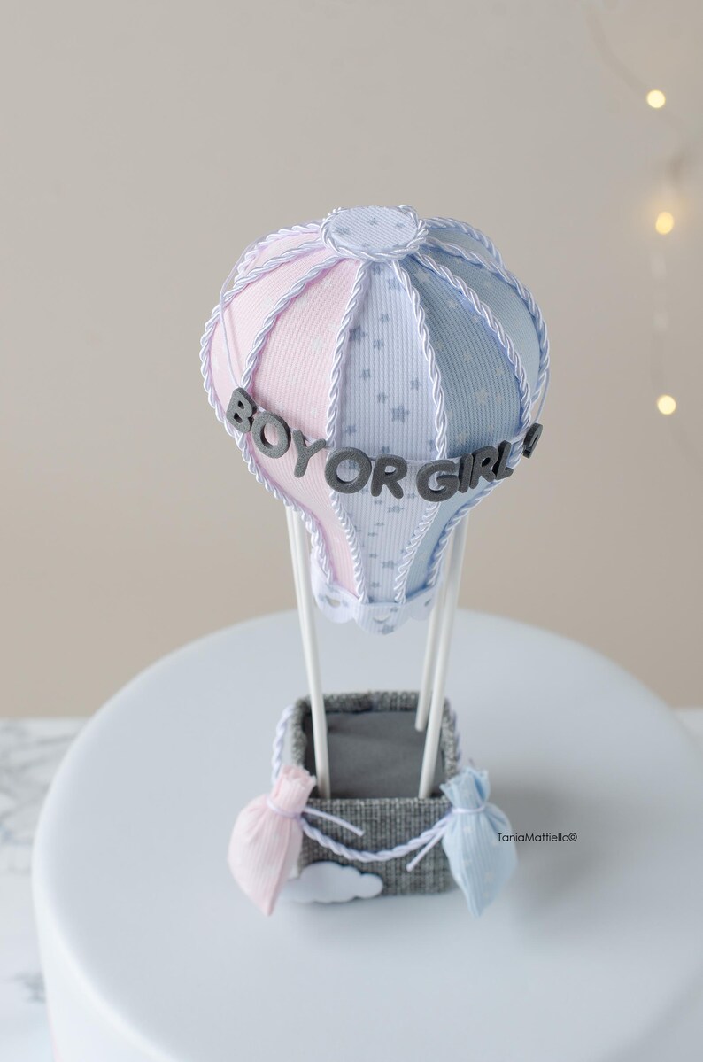 May include: A gender reveal hot air balloon cake topper with a pink and blue striped balloon, white stars, and the text "Boy or Girl!" in gray letters.