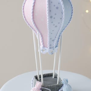 May include: A light blue and pink hot air balloon with white stars and a white rope trim. The balloon is attached to a gray basket with a white rope trim. There are two small pink and blue fabric bags hanging from the basket. The basket is sitting on a white surface.