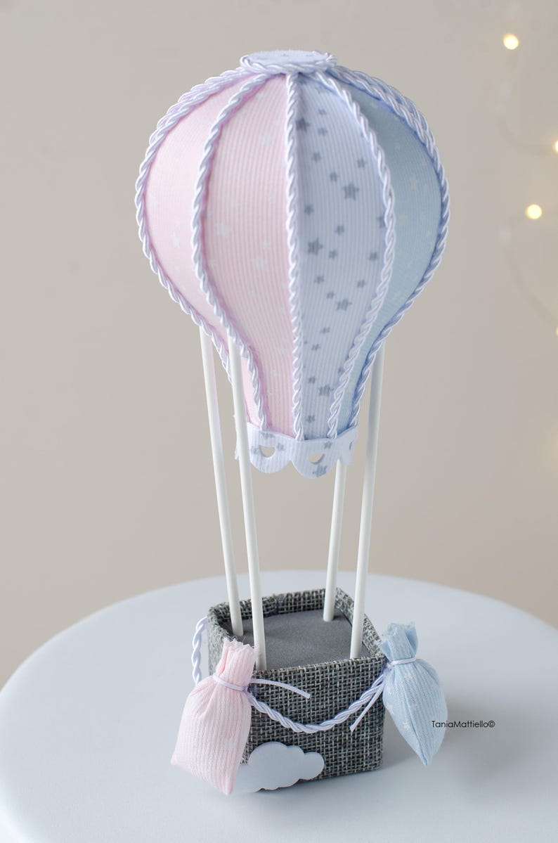 May include: A light blue and pink hot air balloon with white stars and a white rope trim. The balloon is attached to a gray basket with a white rope trim. There are two small pink and blue fabric bags hanging from the basket. The basket is sitting on a white surface.