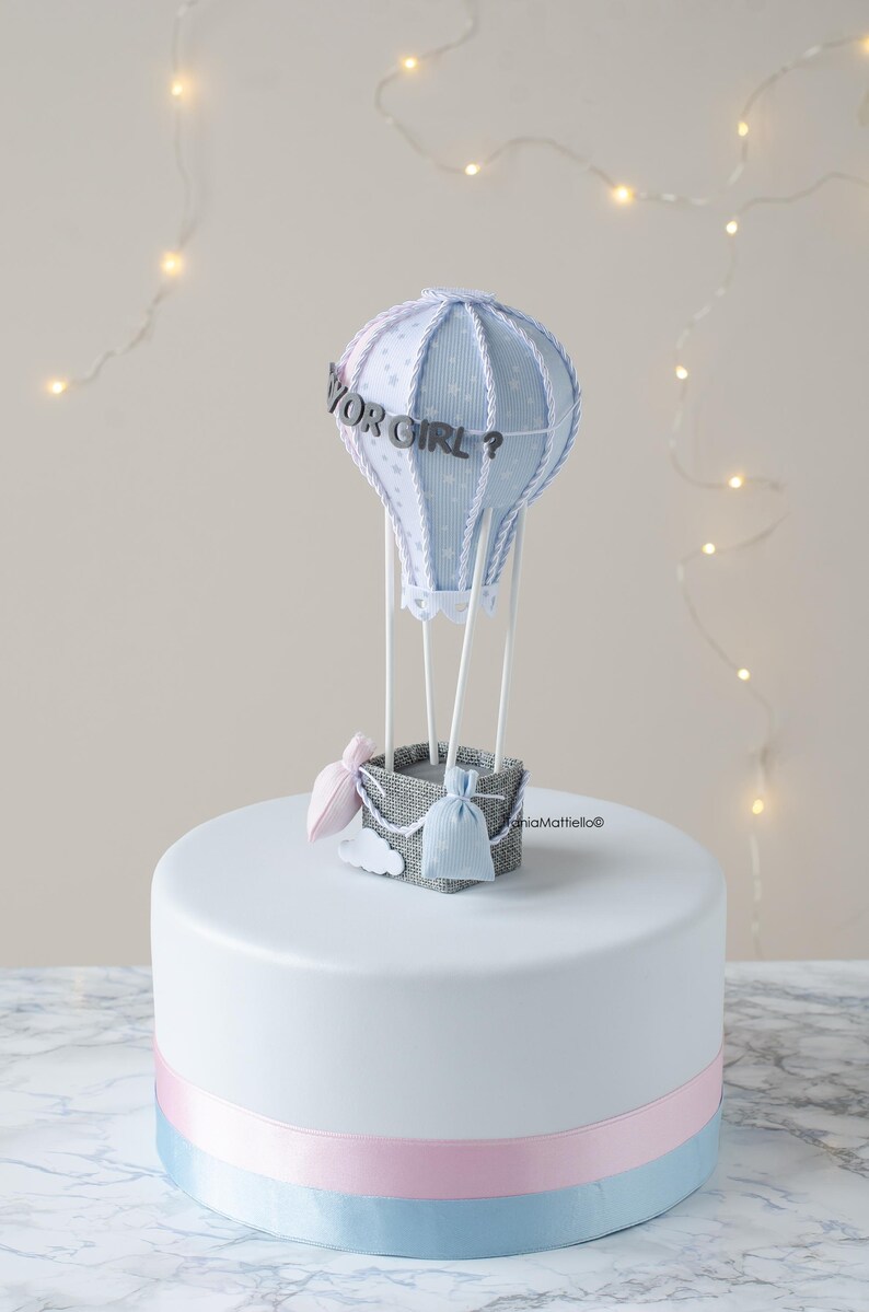 May include: A white cake with a blue and pink ribbon around the bottom. On top of the cake is a blue and white hot air balloon with the words "Boy or Girl?" on it. There are two small bags in the basket of the balloon, one pink and one blue.