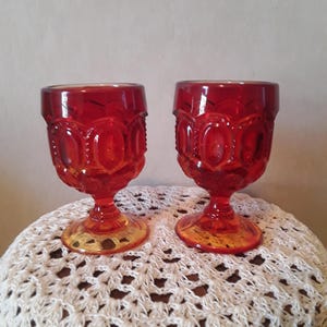 May include: Two vintage red glass goblets with a detailed pattern of moons and stars. The goblets have a short stem and a round base. The glass has a rich, translucent color. The goblets are displayed on a white crocheted surface.