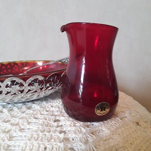 May include: A ruby red glass pitcher with a small spout and a textured surface. A gold sticker with the text "R SWEDEN" is on the side. A decorative silver bowl with a red interior is partially visible.